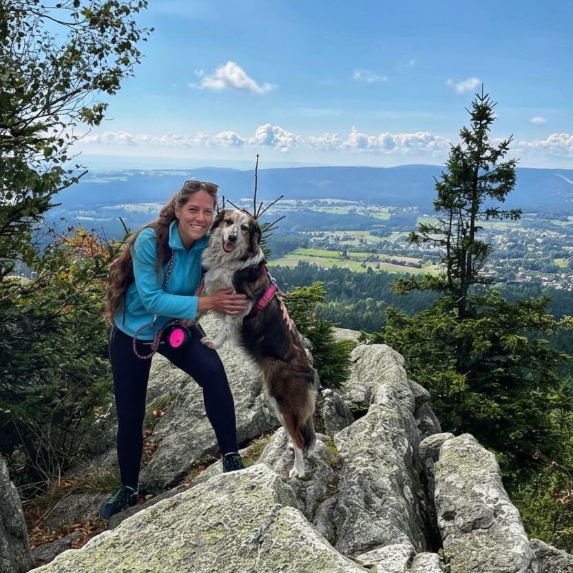 Eine Frau steht mit ihrem Hund auf einem Felsen. Der Himmel ist blau, die Sonne scheint. Der Hund steht mit den Vorderpfoten auf dem Oberschenkel der Frau. Beide sehen glücklich aus.